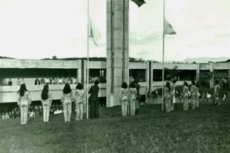 A imagem em sépia mostra uma cerimônia ao ar livre em frente a um edifício de concreto. Em primeiro plano, um grande grupo de pessoas está reunido em uma área gramada, com várias mulheres vestindo calças e blusas claras e um homem de terno escuro, de costas para a câmera, próximo a um mastro alto. Três mastros com bandeiras içadas estão dispostos no centro da composição. Ao fundo, um edifício de concreto aparente com múltiplas aberturas e varandas se estende horizontalmente, com muitas pessoas observando a cerimônia de suas varandas e janelas. O céu está claro e com poucas nuvens, e a iluminação é natural e uniforme.