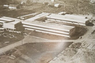 A imagem é uma fotografia aérea em preto e branco de um complexo de edifícios de múltiplos andares, dispostos em formato de "U" ou angular, com longas fachadas e muitas janelas retangulares. Os edifícios parecem ter telhados planos ou levemente inclinados. Em primeiro plano, há uma área gramada com caminhos pavimentados e o que parece ser um lago ou espelho d'água de formato irregular. Um veículo pequeno é visível em uma das vias de acesso aos edifícios. Ao fundo, a paisagem se estende com vegetação e algumas outras construções mais dispersas, indicando um ambiente suburbano ou mais afastado do centro urbano.