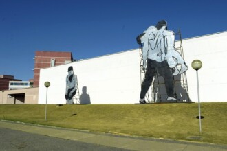 A imagem exibe uma área externa com um gramado verde em primeiro plano, que se eleva em uma pequena colina em direção a uma parede branca e longa. Sobre o gramado, há duas grandes esculturas planas, que representam figuras humanas em preto e branco. A escultura à direita mostra uma figura de pé, carregando algo em seus braços, apoiada por uma estrutura metálica visível atrás dela. A escultura à esquerda retrata uma figura sentada. Entre as esculturas e no gramado, há postes de luz com luminárias esféricas. Ao fundo, atrás da parede branca, aparecem edifícios com fachadas em tons de vermelho e branco, sob um céu azul claro e sem nuvens.