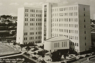 A imagem em sépia mostra um complexo de edifícios de múltiplos andares, predominantemente de cor clara, com estilo arquitetônico que remete a meados do século XX. O edifício principal consiste em duas torres altas e retangulares, com muitas janelas dispostas em fileiras, conectadas por uma estrutura central que parece abrigar escadas ou elevadores. Em frente às torres, há um edifício mais baixo com um telhado arredondado e colunas verticais na fachada. A área em torno dos edifícios é ajardinada, com caminhos e algumas árvores pequenas. Vários carros e alguns ônibus estão estacionados ou circulando nas ruas ao redor e dentro do complexo. Ao fundo, à esquerda, uma encosta com casas pode ser vista.