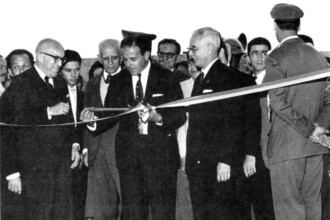 A imagem em preto e branco captura um evento cerimonial de inauguração, com um grupo de homens em destaque. No centro da composição, três homens estão segurando e prestes a cortar uma fita, que possui uma faixa de cor clara e uma faixa escura, estendida horizontalmente. O homem no centro usa um terno escuro, camisa branca e gravata escura, e está segurando uma tesoura para o corte da fita. Ele é ladeado por outros dois homens mais velhos, também de terno e óculos, que o auxiliam. Um deles, à esquerda, sorri e gesticula. Ao redor deles, vários outros homens, alguns em trajes civis e outros com uniformes militares, observam a cena. Um deles, no canto superior direito, usa uma boina. O fundo da imagem é indistinto.