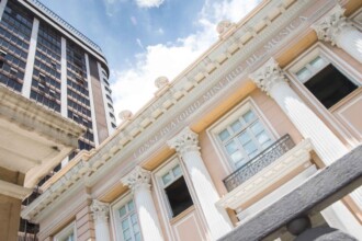 A imagem apresenta a fachada de um edifício histórico em estilo neoclássico, predominantemente na cor pêssego claro com detalhes em branco e dourado. Colunas coríntias decoradas sustentam uma cornija detalhada, sobre a qual está gravado "CONSERVATORIO MINEIRO DE MUSICA" em letras maiúsculas. As janelas retangulares, algumas com sacadas de ferro forjado, estão visíveis. No lado esquerdo da imagem, um arranha-céu moderno, com fachada escura e muitas janelas, eleva-se contrastando com o estilo do Conservatório. O céu acima é azul com algumas nuvens brancas visíveis. O ponto de vista da foto é de baixo para cima, enfatizando a grandiosidade dos edifícios.