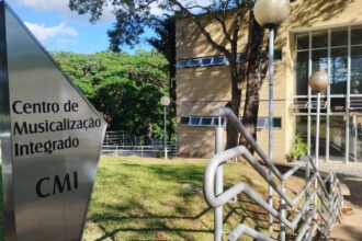 A imagem mostra a entrada de um edifício em um ambiente universitário, em um dia ensolarado. No primeiro plano esquerdo, uma placa metálica triangular exibe o texto "Centro de Musicalização Integrado" e, abaixo, a sigla "CMI", tudo em letras escuras. Ao lado direito da placa, um corrimão de metal cinza acompanha uma escada ou rampa que não está totalmente visível. Ao fundo, um edifício de cor bege com grandes janelas, algumas com brises, se estende. Árvores com folhagem verde densa estão espalhadas pela cena, projetando sombras no gramado verde em frente ao prédio. Postes de iluminação com luminárias esféricas estão presentes no gramado e próximos ao edifício. O céu é azul com algumas nuvens brancas visíveis.