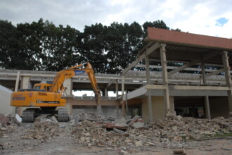 A imagem mostra uma cena de demolição de um edifício de estrutura de concreto. Uma grande escavadeira amarela com uma longa lança equipada com um rompedor hidráulico está em primeiro plano à esquerda, trabalhando na estrutura parcialmente destruída. Há poeira e detritos voando ao redor do ponto de impacto do rompedor. O edifício em demolição tem vários pilares e vigas de concreto expostos, com seções do telhado já removidas ou desabadas. Pilhas de entulho, consistindo principalmente de tijolos quebrados, concreto e outros materiais de construção, estão espalhadas ao redor da base da estrutura. Ao fundo, há uma fileira densa de árvores altas com folhagem escura sob um céu nublado.