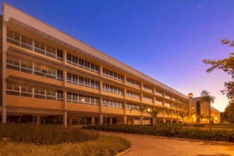 A imagem mostra um grande edifício moderno de múltiplos andares ao entardecer ou amanhecer, sob um céu que varia de azul escuro a tons mais claros perto do horizonte. O edifício tem uma fachada extensa com muitas janelas retangulares iluminadas, sugerindo que as luzes internas estão acesas. A cor da fachada é clara, possivelmente bege ou amarelo pálido. No primeiro plano, há uma área ajardinada com vegetação rasteira e algumas palmeiras esparsas. Um caminho pavimentado sinuoso corta a área ajardinada, levando em direção ao edifício. A iluminação pública, com postes de luz visíveis, contribui para a iluminação da cena, e árvores escuras podem ser vistas na extremidade direita da imagem.