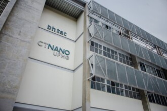 A imagem mostra um edifício moderno, com uma fachada que combina paredes brancas e detalhes em cinza. A parte superior esquerda da construção exibe o logotipo "bh.tec" em letras azul-turquesa e, logo abaixo, "CTNANO UFMG" em um tom de azul similar. O lado direito do edifício possui diversas janelas horizontais e estruturas de brises metálicos na cor cinza, que se estendem por vários andares. Elementos de concreto aparente e metal compõem a arquitetura da edificação. O céu, ao fundo, está nublado, com tons de cinza claro.