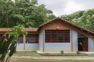 A imagem mostra uma construção de um andar com telhado de telhas cerâmicas em tons de laranja e vermelho. As paredes do edifício são pintadas em uma combinação de azul claro na parte superior e um tom pêssego/laranja na parte inferior, com detalhes em vermelho escuro nas esquadrias das janelas e portas. As janelas são grandes e retangulares, com divisórias internas. Na frente da construção, há uma área de gramado e um caminho de concreto. Plantas com folhas grandes e alongadas, em tons de verde, estão visíveis no canto inferior esquerdo. Ao fundo e nas laterais da construção, há uma densa vegetação de árvores de folhagem verde escura, indicando um ambiente arborizado. O céu, visível no topo da imagem, é claro, com algumas nuvens.