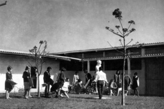 A imagem em preto e branco retrata um pátio externo, provavelmente de uma instituição de ensino de arte, com um grupo de aproximadamente 15 pessoas, a maioria jovens, engajadas em atividades de desenho ou escultura. O pátio é pavimentado ou de terra batida, com algumas árvores jovens e esparsas. No centro do pátio, uma figura feminina, possivelmente uma estátua ou busto, serve de modelo para os estudantes, que estão sentados em cadeiras dobráveis ou no chão, com pranchetas e materiais de desenho. O cenário é delimitado por edifícios baixos com telhados de telhas claras e uma estrutura de apoio em pilares, criando uma área sombreada na parte direita da imagem. O céu é claro e a iluminação geral é forte.