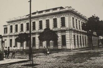 Fotografia em preto e branco da época mostrando o edifício da Escola de Engenharia. A construção ocupa a esquina de duas ruas pavimentadas com paralelepípedos e possui dois andares, com fachada em estilo neoclássico. O prédio é retangular e simétrico, com janelas altas e retangulares dispostas em sequência, todas com molduras decorativas. O andar térreo e o superior são divididos por uma faixa horizontal ornamentada, e o topo da edificação é finalizado com uma platibanda decorativa. Na calçada, há árvores bem podadas e postes de iluminação antigos. Algumas pessoas caminham ou estão paradas na esquina, vestidas com trajes formais da época. À esquerda da imagem, uma placa fixada na parede indica o nome da praça: "Praça Rui Barbosa".