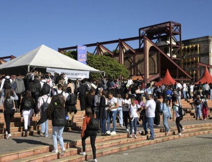 A imagem exibe um ambiente externo movimentado com um grande grupo de estudantes caminhando por uma ampla escadaria e uma praça. A cena central é composta por degraus largos, que levam a uma área pavimentada onde muitas pessoas estão reunidas. No lado esquerdo, uma tenda branca com o topo triangular e a inscrição "MOSTRA ... PRAÇA DE SERVIÇOS" parcialmente visível está montada, e aglomerados de pessoas estão ao seu redor. Ao fundo, estruturas de um edifício com grandes formas geométricas em tons de marrom e vermelho são visíveis, bem como um edifício mais distante com fachada amarela. O céu é azul claro com poucas nuvens, indicando um dia ensolarado.
