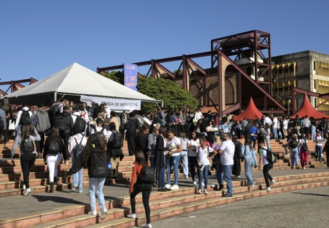 A imagem exibe um ambiente externo movimentado com um grande grupo de estudantes caminhando por uma ampla escadaria e uma praça. A cena central é composta por degraus largos, que levam a uma área pavimentada onde muitas pessoas estão reunidas. No lado esquerdo, uma tenda branca com o topo triangular e a inscrição "MOSTRA ... PRAÇA DE SERVIÇOS" parcialmente visível está montada, e aglomerados de pessoas estão ao seu redor. Ao fundo, estruturas de um edifício com grandes formas geométricas em tons de marrom e vermelho são visíveis, bem como um edifício mais distante com fachada amarela. O céu é azul claro com poucas nuvens, indicando um dia ensolarado.