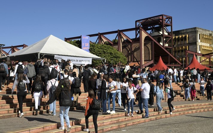 A imagem exibe um ambiente externo movimentado com um grande grupo de estudantes caminhando por uma ampla escadaria e uma praça. A cena central é composta por degraus largos, que levam a uma área pavimentada onde muitas pessoas estão reunidas. No lado esquerdo, uma tenda branca com o topo triangular e a inscrição "MOSTRA ... PRAÇA DE SERVIÇOS" parcialmente visível está montada, e aglomerados de pessoas estão ao seu redor. Ao fundo, estruturas de um edifício com grandes formas geométricas em tons de marrom e vermelho são visíveis, bem como um edifício mais distante com fachada amarela. O céu é azul claro com poucas nuvens, indicando um dia ensolarado.