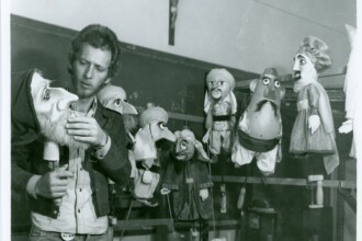 A imagem em preto e branco retrata um homem barbudo, de cabelo encaracolado, vestindo uma camisa de botões clara e calça jeans, interagindo com vários bonecos de fantoches. Ele está no lado esquerdo da imagem, segurando um dos bonecos com as duas mãos, que representa uma figura mais velha com barba e touca. Ao redor do homem, outros cinco bonecos pendurados em estruturas visíveis ou dispostos sobre uma superfície formam uma pequena coleção, cada um com características distintas de vestimenta e fisionomia, variando em tamanho e cor. Um crucifixo está pendurado na parede escura ao fundo, e a iluminação da cena é difusa, vindo principalmente da frente, criando poucas sombras.