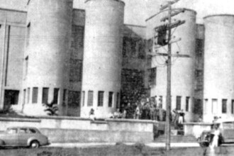 A imagem em preto e branco mostra a fachada de um grande edifício com arquitetura distinta. O prédio é caracterizado por diversas torres cilíndricas que se elevam verticalmente, intercaladas por seções com janelas retangulares. As torres possuem uma série de pequenas aberturas verticais ao longo de sua base, e algumas delas parecem ter janelas maiores nos andares superiores. Em frente ao edifício, no nível da rua, há um muro baixo e uma calçada. No lado esquerdo, um carro de modelo antigo está estacionado. No lado direito, um ônibus também de modelo antigo está parado. Um poste de madeira com fios e isoladores está visível no centro da imagem, um pouco à frente do prédio. No centro da fachada do edifício, em uma área entre duas torres, um grupo de pessoas está reunido na entrada principal ou em uma escadaria, sugerindo atividade no local. O céu não é visível e a iluminação da cena é uniforme.