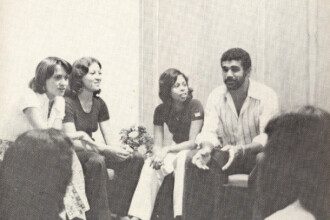 A imagem em preto e branco retrata um grupo de pessoas sentadas em círculo em um ambiente interno, que parece ser uma reunião ou discussão. No centro da parte superior da imagem, quatro indivíduos estão visíveis da cintura para cima, sentados em cadeiras. À esquerda, duas mulheres de cabelo escuro e curtos estão voltadas uma para a outra, conversando. A mulher da esquerda veste uma blusa clara e a da direita uma blusa escura. Ao lado delas, uma terceira mulher, também de cabelo escuro e curto, veste uma blusa escura e calças claras, segurando o que parece ser um pequeno buquê de flores. À direita dela, um homem barbudo, de cabelo escuro e encaracolado, veste uma camisa clara de manga curta e calças escuras, gesticulando com as mãos enquanto fala. Na parte inferior da imagem, as cabeças e ombros de outras pessoas são visíveis, indicando que elas também estão sentadas em um círculo. O fundo é uma parede clara com uma textura sutil. A iluminação é uniforme e suave.