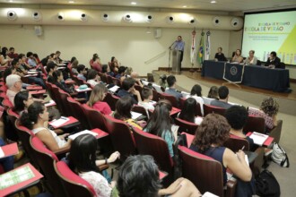 A imagem mostra um auditório grande, repleto de pessoas sentadas em cadeiras estofadas de cor vinho. As fileiras de cadeiras estão dispostas em uma inclinação que leva ao palco, na frente da sala. As pessoas da plateia, a maioria com as costas para a câmera, estão concentradas em uma apresentação. No palco, à direita, há uma mesa com cinco pessoas sentadas, e um banner branco com o texto "Escola de Verão" está ao lado delas. Ao lado, um homem está no púlpito falando ao microfone. O ambiente é iluminado por luzes artificiais e as paredes são de cor clara.