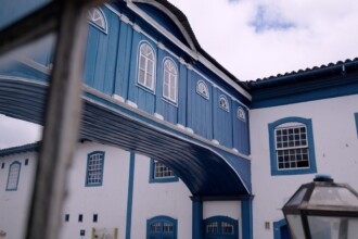 A imagem mostra uma mulher em um ambiente que parece ser um laboratório, com iluminação clara. Ela está sentada de perfil, usando óculos de grau, luvas azuis de proteção e um jaleco branco. A mulher está com a cabeça inclinada para baixo, focada em uma tarefa. Em suas mãos, ela segura uma caneta e um fichário, enquanto o fundo da imagem mostra prateleiras com objetos, caixas de armazenamento e luminárias de mesa.