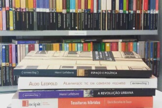 A imagem mostra uma pilha de cinco livros de capa dura e brochura, dispostos na horizontal, em primeiro plano. O livro do topo tem uma capa clara com o título "Espaço e Política" e o nome do autor "Henri Lefebvre", publicado pela Editora UFMG. Abaixo dele, há um livro de capa branca com o título "Almanaque de um Condado Arenoso", de "Aldo Leopold", também com o selo da Editora UFMG. O terceiro livro da pilha é de capa azul escura, com o título "A Revolução Urbana", de "Henri Lefebvre". O quarto livro, de capa vermelha, possui o título "Tessituras híbridas" e o nome da autora "Gisela Barcellos de Souza". O último livro, na base da pilha, tem capa escura e exibe o título "Mais Médicos". Atrás da pilha de livros, há duas prateleiras de uma estante com livros. A prateleira superior mostra uma variedade de livros organizados lado a lado, com capas de cores diversas, como azul, vermelho, laranja, amarelo e preto. A prateleira inferior, um pouco mais baixa, também contém livros enfileirados, com os títulos e autores visíveis em algumas lombadas. As prateleiras são cinzentas.