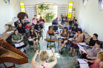 A imagem mostra um grupo de cerca de 20 pessoas em um ambiente interno, que se assemelha a uma sala de ensaio. As pessoas estão sentadas em cadeiras em um semicírculo, cada uma com uma partitura musical à sua frente. No centro, uma mulher de costas para a câmera, com cabelos curtos e claros, está de pé, conduzindo o grupo com as mãos. À sua frente, um cravo de madeira com o tampo aberto e decorado se destaca. Ao fundo, uma porta aberta revela uma paisagem com vegetação, e janelas coloridas em estilo vitral estão nas paredes laterais.
