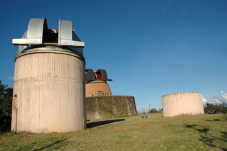 A imagem mostra uma paisagem com três grandes estruturas cilíndricas ao ar livre, sob um céu azul claro. Em primeiro plano, uma estrutura de cor clara com uma cúpula de metal aberta no topo domina o lado esquerdo da imagem. Ao fundo, outras duas estruturas semelhantes são visíveis, uma com uma cúpula metálica, e a outra de cor mais escura e com uma construção circular de pedra e musgo ao redor. A grama verde cobre o chão, e arbustos e vegetação rasteira são visíveis nos arredores.