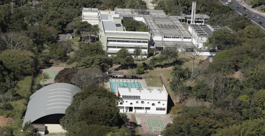 A imagem é uma fotografia aérea de uma área arborizada, que abriga diversos edifícios e instalações esportivas. No centro, um grande complexo de prédios brancos de dois andares com telhados planos se destaca, cercado por uma densa vegetação. As janelas dos edifícios são retangulares e há uma torre de água ou chaminé no telhado. À frente do complexo principal, um prédio branco de formato alongado com janelas circulares está localizado próximo a uma piscina retangular de água azul. Ao lado da piscina, há duas quadras de tênis de saibro. Na parte inferior esquerda da imagem, um ginásio com um telhado de metal arqueado é visível. Uma estrada com tráfego de veículos percorre a parte superior direita da foto.