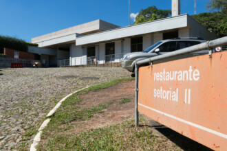 A imagem mostra um edifício de fachada branca, com um telhado de dois andares, sob um céu azul e com nuvens brancas. Na frente do edifício, uma placa de cor laranja com letras brancas informa "restaurante setorial II". O edifício tem uma rampa de acesso, portas de vidro e janelas retangulares. Um carro cinza está estacionado à direita, e a rua, em primeiro plano, é feita de paralelepípedos.