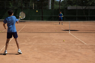 A imagem mostra um jovem em uma quadra de tênis de saibro, de costas para a câmera, se preparando para rebater uma bola. Ele veste uma camisa azul e shorts escuros. Uma raquete de tênis está em suas mãos, e uma bola é visível no ar, prestes a ser atingida. Outro jogador está do outro lado da quadra, atrás da rede, em uma posição de espera. A quadra tem a marcação em branco, e algumas bolas estão no chão. O fundo da quadra é cercado por árvores e uma cerca de metal.