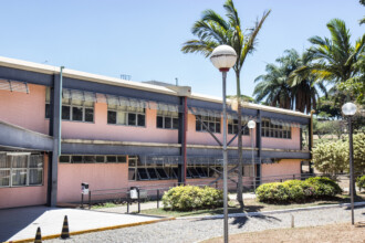 A imagem mostra a vista externa de um edifício de dois andares com paredes de cor salmão e uma faixa azul escura na altura do telhado. O prédio tem grandes janelas com persianas horizontais. Há uma rampa de acesso com corrimão de metal na frente do prédio. Em primeiro plano, o chão é pavimentado com blocos de concreto e pedras, formando uma passarela. Há árvores e palmeiras altas, assim como arbustos baixos e redondos na frente do prédio. Um poste de iluminação com duas luminárias brancas está no centro da imagem. O céu está limpo, com poucas nuvens, e a luz do sol é intensa, criando sombras no chão.