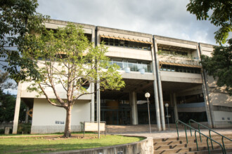 A imagem mostra um edifício de três andares com fachada de concreto, janelas retangulares e varandas com grades. Uma escadaria de concreto com corrimões verdes leva à entrada principal. No primeiro plano, uma grande árvore com galhos esparsos se destaca, e um gramado verde está visível na parte inferior. O fundo da imagem é preenchido por um céu nublado, e o edifício é emoldurado por outras árvores e folhagens.