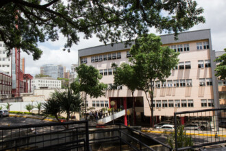 A imagem mostra uma vista externa de um edifício de cor salmão, com quatro andares e muitas janelas, em um dia claro com algumas nuvens no céu. A entrada principal do prédio é acessada por uma escada e uma rampa branca, visíveis na parte inferior da imagem. Ao redor da entrada, há várias pessoas caminhando ou paradas, e algumas árvores com folhagem verde. No primeiro plano, uma grade de metal preta delimita uma área elevada e sombreada, com uma árvore grande e frondosa no topo. A parte inferior da imagem mostra um estacionamento com alguns carros brancos. Ao fundo, uma paisagem urbana é visível, com outros edifícios altos, inclusive um prédio vermelho e branco à esquerda e arranha-céus cinzentos ao longe.