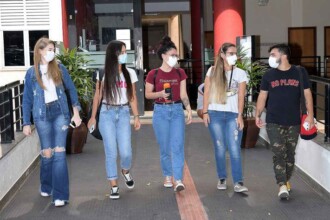 A imagem mostra cinco jovens, três mulheres e dois homens, caminhando juntos em um corredor coberto. Todos estão usando máscaras de proteção facial brancas. O grupo está voltado para a esquerda. A mulher à frente, da esquerda, tem cabelo loiro e usa uma jaqueta jeans azul, calça jeans e tênis brancos. A segunda mulher, da esquerda, tem cabelo escuro e comprido, e usa uma camiseta branca, calça jeans e tênis preto. A mulher do centro, de cabelo castanho cacheado, usa uma camiseta vermelha escura e calça jeans. As outras duas pessoas, um homem e uma mulher, usam jeans e camisetas escuras. O ambiente é iluminado pela luz do sol, e plantas em vasos são visíveis no canto esquerdo.