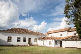 A imagem mostra um conjunto de edifícios antigos de paredes brancas com telhados de telhas de barro, em um ambiente externo. No lado esquerdo, um prédio com janelas de madeira retangulares, com grades, e uma porta central pode ser visto. No centro, uma área de grama verde se estende em frente aos edifícios. À direita, outro prédio branco, com telhado de telhas de barro e uma pequena cruz no topo, é visível. Pequenas janelas retangulares, com caixilhos azuis, são vistas na fachada. A iluminação é natural, e o céu, no plano superior, é azul com nuvens brancas e grandes. Uma parte de uma árvore é visível no canto superior direito.