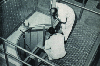 A imagem em preto e branco mostra dois homens em um ambiente industrial, de um ponto de vista superior. Os dois homens, vestidos com macacões brancos, estão agachados e trabalhando em uma área circular com um poço. Eles estão cercados por um piso de grade de metal e corrimões de segurança. O ambiente tem uma iluminação forte, e o chão parece ser de concreto, com uma parede de tijolos à vista ao fundo.