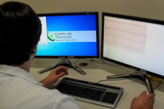 A imagem mostra a visão das costas de uma pessoa sentada em frente a dois monitores de computador. A pessoa veste um jaleco branco e usa um teclado e um mouse. Os monitores estão lado a lado sobre uma mesa de cor clara. O monitor da esquerda exibe a tela inicial de um sistema ou programa, com um fundo azul e uma faixa branca. O logotipo do "Centro de Telessaúde" e a inscrição "Hospital das Clínicas - UFMG" são visíveis. O monitor da direita exibe um gráfico de eletrocardiograma (ECG), mostrando uma série de linhas e picos pretos sobre um fundo branco.