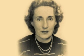 A imagem é um retrato em preto e branco de uma mulher, com uma leve tonalidade sépia. Ela está olhando diretamente para a câmera, com os cabelos ondulados e curtos na altura dos ombros. A mulher veste uma blusa escura com listras horizontais e usa um colar de duas voltas de pérolas. A fotografia tem um fundo liso e claro, com as bordas desfocadas.