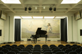 A imagem mostra o interior de um auditório, com um palco de madeira em primeiro plano. Um piano de cauda preto com um banquinho está posicionado no centro do palco. Ao fundo, há uma grande tapeçaria ou mural com uma pintura que representa duas figuras humanas, uma em pé à esquerda e outra ajoelhada à direita. Cortinas pretas emolduram o palco e, à frente dele, várias fileiras de cadeiras estofadas escuras estão dispostas.