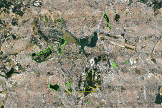 A imagem é uma fotografia aérea de uma área urbana densamente construída, vista de cima. A paisagem é composta por um vasto tecido urbano de construções, com ruas e avenidas que se cruzam. No centro da imagem, há um grande lago de cor verde-escura. Várias áreas de vegetação, em formato irregular, estão contornadas por linhas verdes, e duas delas são destacadas em amarelo, com a inscrição "Estação Ecológica UFMG". No canto superior direito, uma pista de aeroporto é visível. A iluminação é natural e o céu é claro.