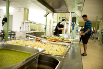 A imagem mostra a área de self-service de um restaurante. Em primeiro plano, bandejas de metal repletas de comida estão dispostas em um balcão. À esquerda, uma bandeja contém um caldo verde. No centro, uma bandeja grande está cheia de salada de repolho e cenoura ralados, com grandes conchas de metal por cima. Ao fundo, outras bandejas de comida são visíveis, e há algumas pessoas servindo-se. Um homem de camisa xadrez azul e bermuda preta está em pé, no lado direito, se servindo de comida. Outros funcionários do restaurante podem ser vistos em segundo plano. O ambiente tem paredes claras e é iluminado por luzes artificiais.
