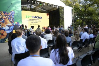 A imagem mostra uma cerimônia em um palco ao ar livre, com uma plateia sentada em cadeiras pretas. Em primeiro plano, a visão é da parte de trás da plateia, onde as pessoas estão sentadas, com as costas para a câmera, observando o palco. O palco tem um telão que exibe a inscrição "59th IChO" e "International Mendeleev Chemistry Olympiad", com a localização "Brazil, Belo Horizonte". No palco, várias pessoas estão em pé. A plateia está sentada em cadeiras de plástico, e uma linha de árvores e folhagem é visível no fundo. A iluminação é natural, proveniente do sol.