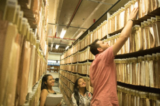 A imagem mostra três pessoas em um corredor estreito de um arquivo ou biblioteca, com prateleiras altas e cheias de pastas suspensas. As pastas são de cor creme e estão dispostas em longas fileiras em ambos os lados do corredor, do chão até o teto. As prateleiras estão numeradas e organizadas por categorias. A iluminação é fornecida por luminárias fluorescentes no teto. Em primeiro plano, à direita, um homem de camisa rosa clara, de pé, alcança uma pasta na prateleira superior. Duas mulheres estão ao lado dele, observando-o. A mulher da esquerda segura um conjunto de papéis em uma pasta branca, e a mulher à sua direita está olhando para o homem com as mãos juntas.