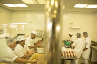 A imagem mostra uma cozinha industrial, com iluminação clara, onde seis pessoas estão trabalhando. Todos usam toucas de cabelo e aventais brancos, e alguns também usam luvas de proteção. As pessoas estão agrupadas em duas bancadas de trabalho. A bancada da esquerda mostra três pessoas manuseando uma grande quantidade de massa marrom em uma tigela amarela. Na bancada da direita, outras duas pessoas parecem estar embalando um produto.