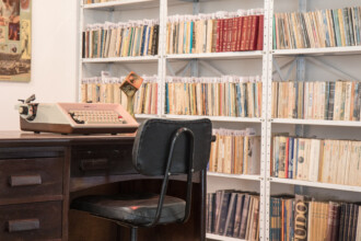 A imagem mostra uma escrivaninha de madeira escura com uma máquina de escrever antiga e uma cadeira à frente. A máquina de escrever é de cor salmão e branca, e está posicionada no lado esquerdo da mesa. Ao lado dela, há uma pequena escultura de metal, de cor laranja e preta. A escrivaninha tem gavetas à esquerda e um pequeno nicho ao centro. A cadeira é preta, com um encosto baixo de metal e um assento estofado, também preto. Ao fundo, há uma grande estante branca com prateleiras cheias de livros. Os livros estão organizados lado a lado, preenchendo completamente as prateleiras. As capas e lombadas têm uma variedade de cores, incluindo vermelho, azul, amarelo e branco. Ao lado esquerdo da estante, uma parte de um quadro é visível, mostrando o que parece ser um carro antigo. O ambiente é iluminado e as paredes são brancas.