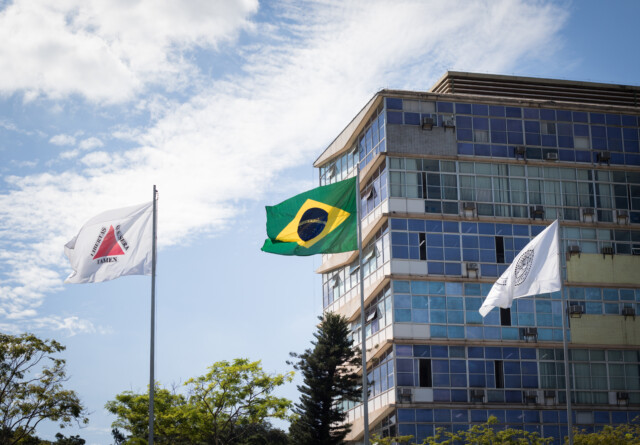 La photo montre un plan en contre-plongée d'un bâtiment et de trois drapeaux sur fond de ciel bleu nuageux. Le drapeau national brésilien, vert, jaune et bleu, flotte au centre. À sa gauche, un autre drapeau blanc avec un triangle rouge et l'inscription « LIBERTAS QUAE SERA TAMEN » est visible. À droite du drapeau brésilien, un troisième drapeau blanc, partiellement masqué, arbore le sceau circulaire de l'Université fédérale du Minas Gerais (UFMG). Les drapeaux flottent sur des mâts devant un bâtiment moderne à la façade composée de panneaux rectangulaires bleus et gris, dont de nombreuses fenêtres. Le bâtiment semble être un immeuble de bureaux ou un bâtiment institutionnel de plusieurs étages. Des branches d'arbres sont visibles en bas de l'image, suggérant un environnement de parc ou de campus. L'éclairage est intense, signe d'une journée ensoleillée.