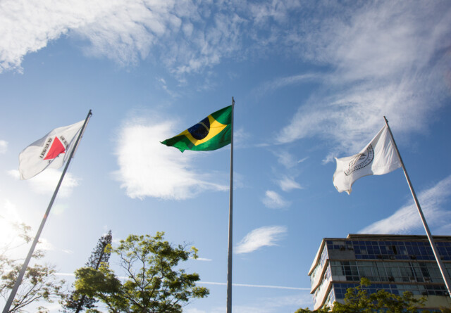 Essa imagem mostra um plano de baixo ângulo que mostra três bandeiras em hastes de metal contra um céu azul com nuvens brancas e esparsas. Da esquerda para a direita, as bandeiras são: a bandeira de Minas Gerais (branca com um triângulo vermelho e o lema "LIBERTAS QUAE SERA TAMEN"), a bandeira nacional do Brasil (verde, amarela e azul) e a bandeira da UFMG (branca com o selo da universidade).  As bandeiras estão tremulando levemente ao vento. O sol está brilhando fortemente, com um reflexo de lente visível no canto superior esquerdo da imagem, o que cria uma luz brilhante e dramática. No canto inferior esquerdo, há a copa de uma árvore, e no canto inferior direito, parte de um edifício moderno e multiandares com painéis de vidro. A perspectiva de baixo para cima faz as bandeiras e o edifício parecerem imponentes.