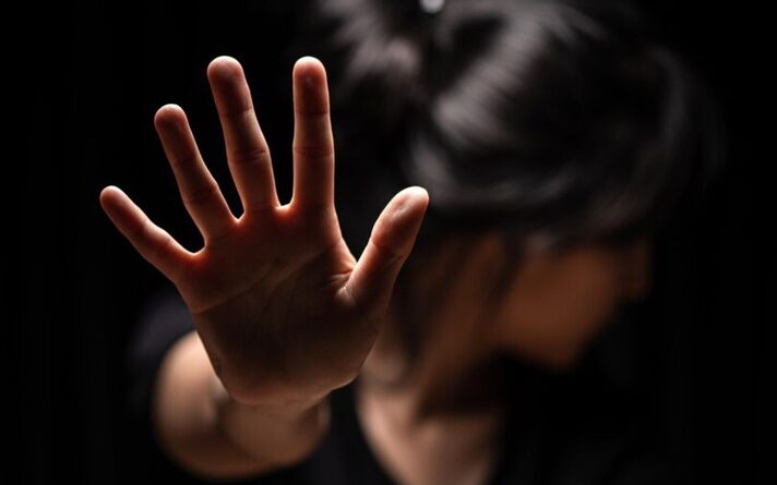 Mulher com a mão levantada em direção à câmera, em um gesto de "pare", com o rosto virado e desfocado ao fundo