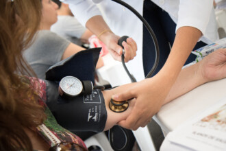A imagem mostra um prédio branco com telhado azul e branco, em uma área com calçamento de pedra e vegetação. Há um arco azul escuro que liga as duas partes do edifício, e o céu, com nuvens brancas e esparsas, está visível no topo da imagem. A parte inferior do arco revela uma vista de um vilarejo com casas de telhado marrom. Em primeiro plano, uma luminária de rua antiga com uma estrutura de ferro escuro se destaca.