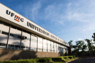A imagem mostra uma vista externa de um edifício de dois andares com uma fachada de cor clara. A parte superior da fachada exibe, em letras grandes e escuras, a sigla "UFMG" e o nome completo "UNIVERSIDADE FEDERAL DE MINAS GERAIS". A parte inferior da fachada é coberta por um revestimento que se assemelha a bambu, e janelas retangulares com ar-condicionado de parede estão no andar de cima. Em frente ao prédio, um jardim bem cuidado com grama e arbustos baixos, com um caminho de terra ao lado, é visível. Ao fundo, a vegetação e as árvores do campus podem ser vistas. O céu, no plano superior, está azul com nuvens brancas, e a luz do sol é intensa, criando reflexos nas janelas.