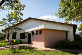 A imagem mostra uma vista externa de um edifício de um andar, com um telhado de telhas vermelhas e uma estrutura principal de cor clara. A entrada do prédio é coberta por uma pequena marquise e tem paredes em um tom de salmão. Na fachada, uma fileira de janelas pequenas, de formato retangular, é visível. Em frente ao prédio, um caminho pavimentado leva à entrada. O edifício está cercado por grama verde, arbustos e árvores com folhas. Na parte da frente, quatro lixeiras coloridas em azul, verde, amarelo e vermelho estão lado a lado. Um poste de iluminação, com duas luminárias, está próximo da entrada. O céu, visível no plano superior, está azul com nuvens brancas.