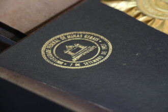 A imagem em close-up mostra um objeto de cor escura, que parece ser a tampa de uma caixa ou capa de um livro. No centro, há um selo circular dourado com a inscrição "UNIVERSIDADE FEDERAL DE MINAS GERAIS" e "7 DE SETEMBRO DE 1927" ao redor de um desenho de um edifício com o lema "INSCIPIT VITA NOVA". A superfície do objeto tem uma textura sutil e o selo dourado está ligeiramente em relevo. A iluminação destaca a textura da superfície, e o fundo da imagem é borrado, sugerindo que o objeto está dentro de uma caixa, com a tampa aberta.