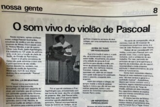 A imagem mostra uma página de jornal em preto e branco, com um título grande no topo da página que diz "O som vivo do violão de Pascoal". O texto da matéria está dividido em várias colunas e se refere a um músico chamado Pascoal Guimarães. No centro da página, uma fotografia em preto e branco mostra um homem, presumivelmente Pascoal, sentado em uma cadeira tocando violão. Ele está vestido com uma camisa de manga curta de cor clara, calças escuras e sapatos. Ao lado da foto, há legendas e textos sobre sua biografia.